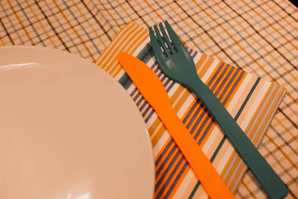 An empty white plate stands on checkered tablecloth colorful cutlery rests, napkin. Table setting top view flatly. Plastic knife and fork in retro vintage style. Kitchen set, disposable cutlery.