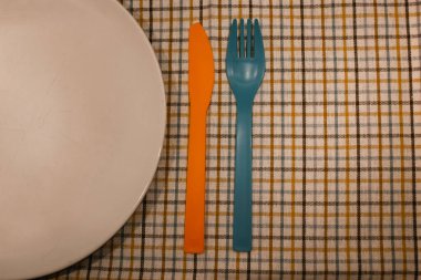 An empty white plate stands on checkered tablecloth colorful cutlery rests, napkin. Table setting top view flatly. Plastic knife and fork in retro vintage style. Kitchen set, disposable cutlery.