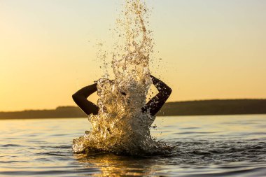 Faceless person in calm water, wave covers person with head. Diving under water on sunset. Dangerous situation on coast. Splashing water in the air. Resting on the shore, beach. Artistic photography.