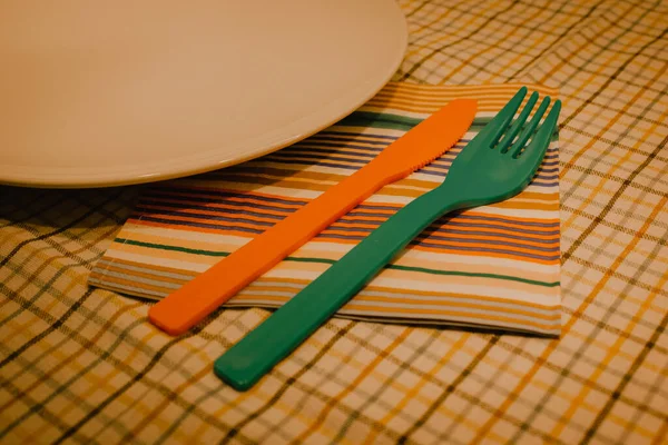 An empty white plate stands on checkered tablecloth colorful cutlery rests, napkin. Table setting top view flatly. Plastic knife and fork in retro vintage style. Kitchen set, disposable cutlery.