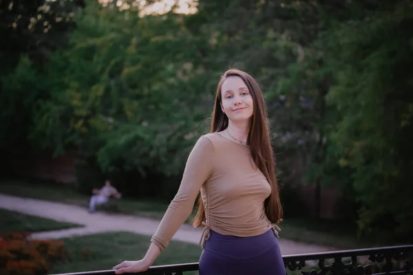 Beautiful white-skinned young woman with long brown hair in tight-fitting casual sportswear standing in a city park in nature. Natural female beauty. Sporty sexy relaxed girl smiling looking in camera