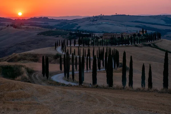 Τοσκάνη, Κρήτη Senesi αγροτικό τοπίο ηλιοβασίλεμα. Εξοχή αγρόκτημα, κυπαρίσσια δέντρα, πράσινο πεδίο, φως του ήλιου και σύννεφο. Ιταλία, Ευρώπη, Καλοκαιρινό αγροτικό τοπίο με καμπύλο δρόμο στην Τοσκάνη, Ιταλία, Ευρώπη — Φωτογραφία Αρχείου