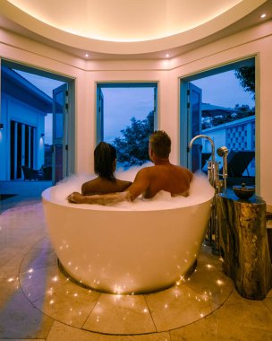 Lüks tatil beldesinde tatilde olan orta yaşlı bir çift küvette oturmuş okyanus manzaralı Avrupalı bir adam ve Asyalı bir kadın küvette oturuyor.