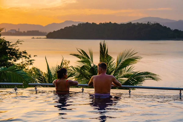 Casal homem e mulher de meia idade na piscina em umas férias de luxo na Tailândia, homens e mulher asiática na piscina olhando para fora sobre a baía assistindo pôr do sol — Fotografia de Stock