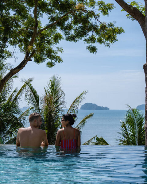 couple man and woman mid age in swimming pool on a luxury vacation in Thailand, men and Asian woman in pool looking out over the bay watching sunset