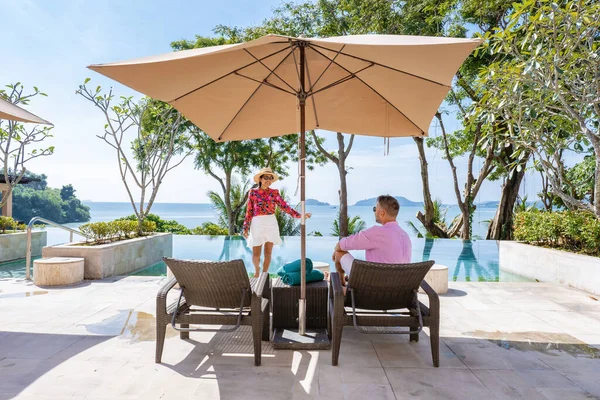 Casal homem e mulher de meia idade na piscina em umas férias de luxo na Tailândia, homens e mulher asiática na piscina olhando para fora sobre a baía assistindo pôr do sol — Fotografia de Stock