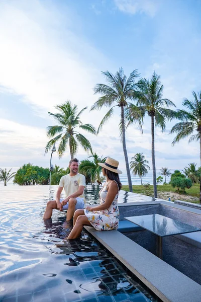 Casal assistindo pôr do sol na piscina infinito em umas férias de luxo na Tailândia, homem e mulher assistindo pôr do sol na borda de uma piscina na Tailândia — Fotografia de Stock