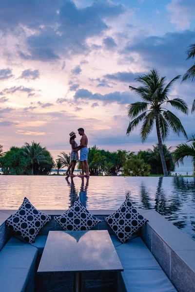 Casal assistindo pôr do sol na piscina infinito em umas férias de luxo na Tailândia, homem e mulher assistindo pôr do sol na borda de uma piscina na Tailândia — Fotografia de Stock