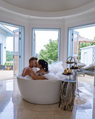 Lüks tatil beldesinde tatilde olan orta yaşlı bir çift küvette oturmuş okyanus manzaralı Avrupalı bir adam ve Asyalı bir kadın küvette oturuyor.