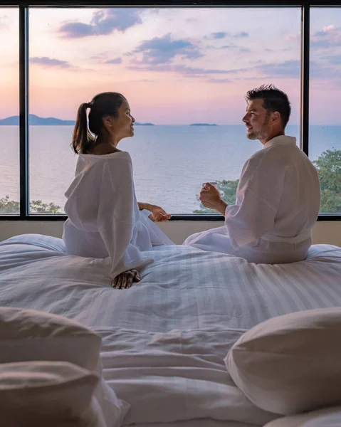 Couple homme et femme en fron de lit dans la chambre au lever du soleil boire une tasse de café, homme européen et femme asiatique dans la chambre d'hôtel pendant le coucher du soleil — Photo