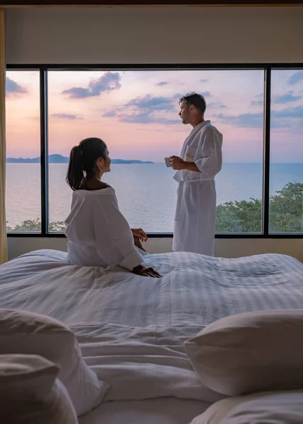 Pareja hombre y mujer en fron de la cama en el dormitorio durante la salida del sol beber una taza de café, hombre europeo y mujer asiática en la habitación de hotel durante la puesta del sol —  Fotos de Stock