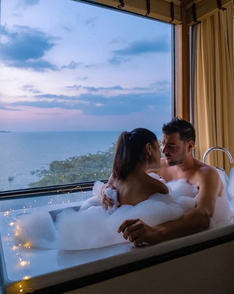 Couple homme et femme dans la salle de bain prendre un bain pendant le lever du soleil, homme européen et femme asiatique dans la chambre d'hôtel pendant le coucher du soleil — Photo