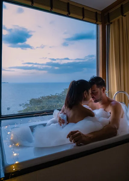 Pareja hombre y mujer en baño tomando un baño durante el amanecer, hombre europeo y mujer asiática en habitación de hotel durante el atardecer —  Fotos de Stock