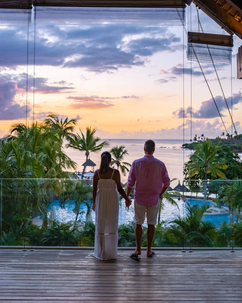 Casal homem e mulher assistindo pôr do sol por uma piscina com palmeiras tropicais durante umas férias de luxo — Fotografia de Stock