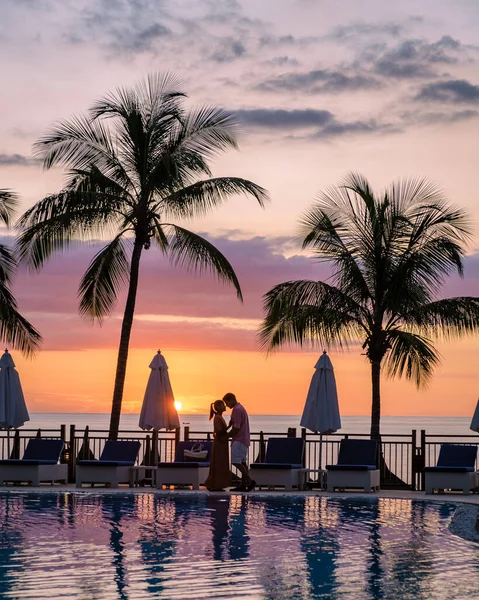 Casal homem e mulher assistindo pôr do sol por uma piscina com palmeiras tropicais durante umas férias de luxo — Fotografia de Stock
