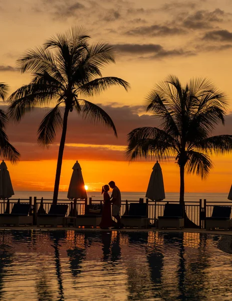 couple man and woman watching sunset by a swimming pool with tropical palm trees during a luxury vacation holiday