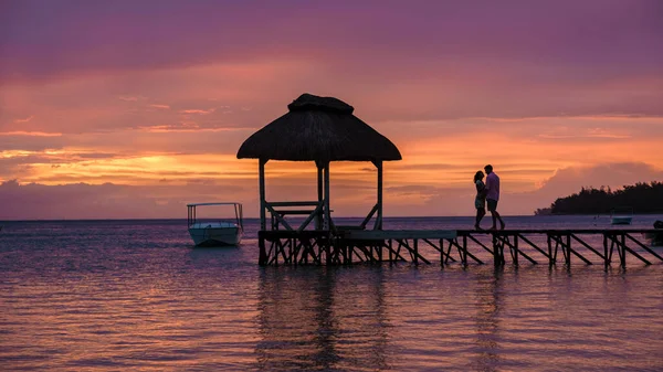 Casal assistindo pôr do sol em uma praia tropical com cais de madeira no oceano, homens e mulheres pôr do sol na praia durante as férias de luxo — Fotografia de Stock