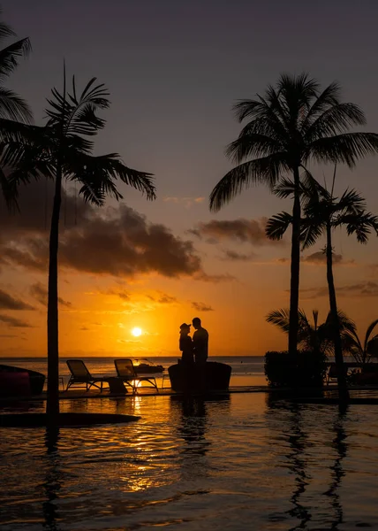 Pár férfi és nő nézi naplementét Mauritius trópusi strandján pálmafákkal a medence mellett, Trópusi naplemente Mauritius strandján — Stock Fotó