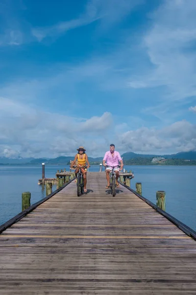 men and woman on a bicycle, a couple of men and woman on a tropical island with a wooden pier jetty in Thailand Phuket Naka Island.