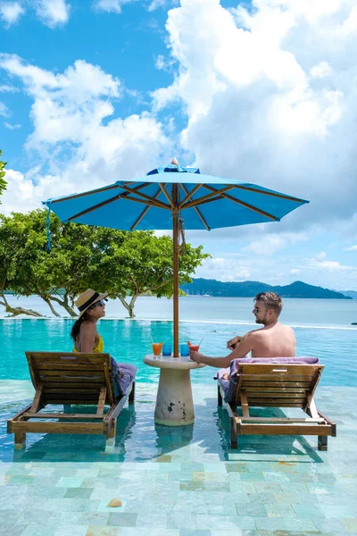 Tayland 'da sonsuz havuzda, okyanusa bakan bir çift Avrupalı erkek ve Asyalı bir kadın Tayland' da lüks bir tatil. Phuket, plaj sandalyeli havuz ve mavi okyanuslu şemsiye.