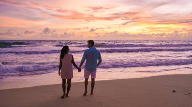 Phuket Tayland sahilinde gün batımı, kumsalda muson yağmurları sırasında renkli günbatımı. Asyalı kadın ve beyaz erkek gün batımını izliyor
