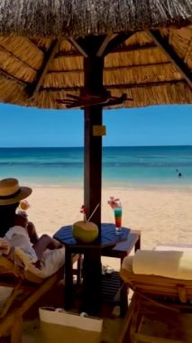 Sahilde şemsiyenin altında, Mauritius 'ta kokteyl içen Asyalı bir kadın. Sahildeki kumsal yatağında mavi bir okyanus var.