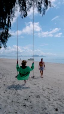Phuket Thailand Naka Island, a couple of men, and women at a swing on the beach. Tropical beach with a swing. Asian women and European men on the beach with swim short