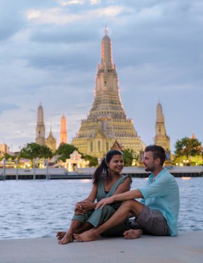 Wat Arun tapınağı Bangkok Tayland, Şafak Tapınağı, Budist tapınağı Chao Phraya Nehri 'nin yanında. Güzel Wat Arun akşam üstü, çift Asyalı kadın ve Avrupalı erkek gün batımını izliyor.