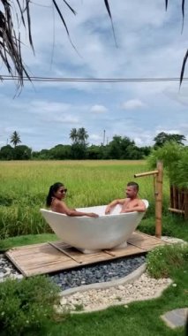 Couple of men and women visiting an eco farm on vacation relaxing in a white bathtub between a beautiful green paddy field in Thailand, bath tub at a home stay farm at the rice field. 