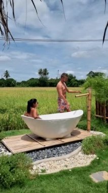 Couple of men and women visiting an eco farm on vacation relaxing in a white bathtub between a beautiful green paddy field in Thailand, bath tub at a home stay farm at the rice field. 