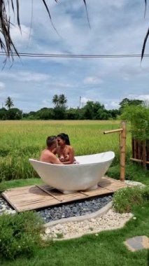 Couple of men and women visiting an eco farm on vacation relaxing in a white bathtub between a beautiful green paddy field in Thailand, bath tub at a home stay farm at the rice field. 
