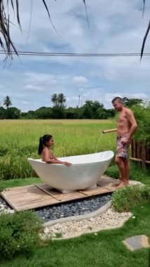 A couple of men and women visiting an eco farm on vacation relaxing in a white bathtub between a beautiful green paddy field in Thailand, and bath tub at a home stay farm at the rice field Thailand