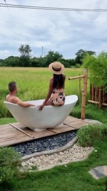 A couple of men and women visiting an eco farm on vacation relaxing in a white bathtub between a beautiful green paddy field in Thailand, and bath tub at a home stay farm at the rice field Thailand