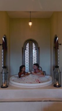 Asian women and European men relax in a bathtub during vacation, a luxury holiday. Couple men and woman drinking wine in the bathtub during a vacation in a luxury resort