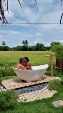 a couple of men and women in a white bathtub outside looking out over a green rice field, men and women in bathtub outside on vacation at a homestay in Thailand with a green rice paddy field