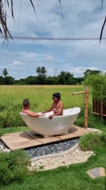 a couple of men and women in a white bathtub outside looking out over a green rice field, men and women in bathtub outside on vacation at a homestay in Thailand with a green rice paddy field