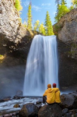 Beautiful waterfall in Canada, couple visit Helmcken Falls, the most famous waterfall in Wells Gray Provincial Park in British Columbia, Canada. couple sitting on a rock looking at the waterfall