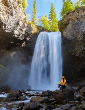 Moul waterfall in Canada, couple visit Moul Falls, the most famous waterfall in Wells Gray Provincial Park in British Columbia, Canada. Asian woman with a yellow sweater and hat looking at a waterfall