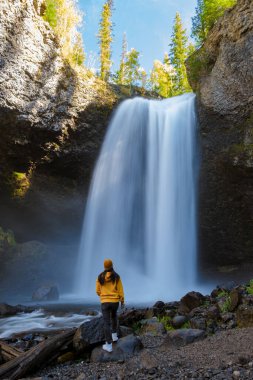 Moul waterfall in Canada, couple visit Moul Falls, the most famous waterfall in Wells Gray Provincial Park in British Columbia, Canada. Asian woman with a yellow sweater and hat looking at a waterfall