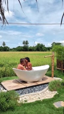 couple men and women in a white bathtub outside looking out over green rice field, couple men and women on vacation at a homestay in Thailand in a bath tub with green rice paddy field