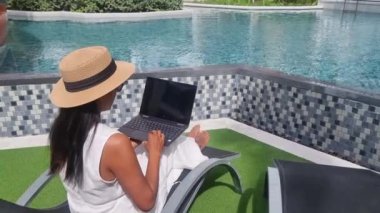 Asian women with hat checking laptop on holiday vacation, digital nomad concept. women with a laptop computer by the pool