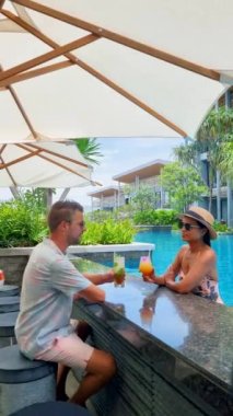 A couple of men and women drinking cocktails by the bar of a tropical pool during vacation. Asian women and caucasian men drinking cocktails during a holiday in a luxury resort