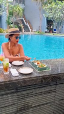 Asian women drinking cocktails by the bar of a tropical pool during vacation. Asian women with hat drinking cocktails during a holiday in a luxury resort