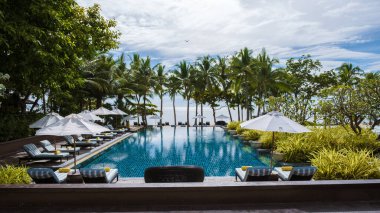 Lüks bir tatil köyünde palmiye ağaçları ve plaj sandalyeleri olan tropik bir havuz, okyanusa bakan palmiye ağaçları olan bir havuz. Havuzda plaj havlusu olan plaj sandalyeleri