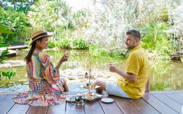 Afternoon tea at a water pond, high tea in a tropical garden in Thailand. Couple of men and women having high tea in a tropical garden