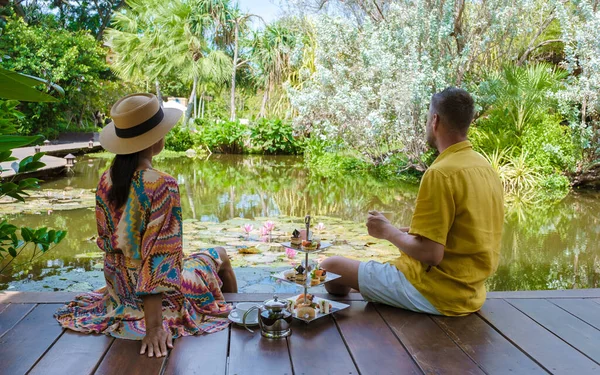 Afternoon tea at a water pond, high tea in a tropical garden in Thailand. Couple of men and women having high tea in a tropical garden
