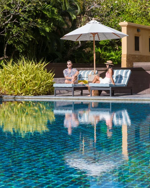 a couple of men and women relaxing by a swimming pool on a beach bed with reflections in the pool. couple on vacation relaxing on a chair by the pool