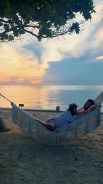 Hua Hin Tayland 'da tropik bir plajda güneşin doğuşunu izleyen bir çift kadın ve erkek. Huahin Tayland sahilindeki hamakta Asyalı kadınlar ve Avrupalı erkekler