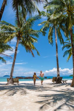 Tayland 'da beyaz tropik bir plajda yürüyen yüzme şortlu erkekler ve kadınlar, Koh Kham Trat. Erkekler kısa yüzer ve Asyalı kadınlar sahilde palmiye ağaçları ve beyaz kumlarla