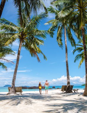 Koh Kham Trat Tayland, beyaz tropik bir plajda yüzme elbiseli palmiye ağaçlarıyla yürüyen birkaç kadın ve erkek.